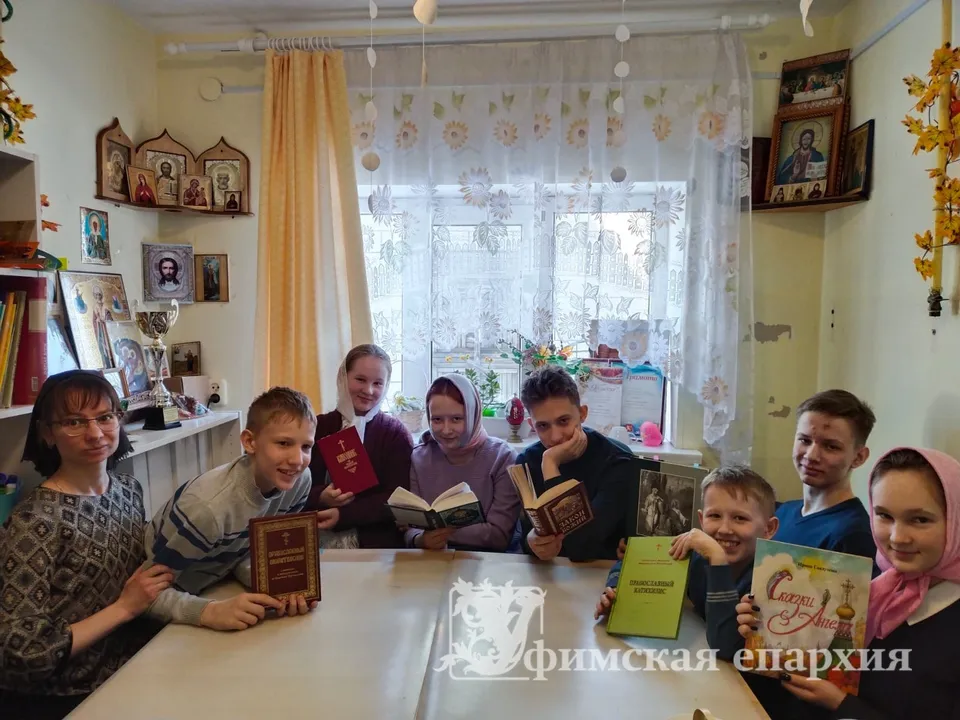 Учительница и восемь детей сидят за столом с книгами в классной комнате с иконами